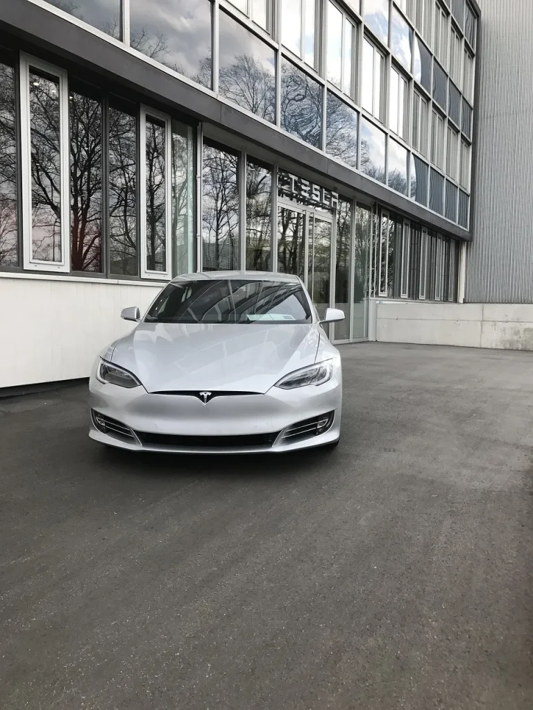 Das Foto zeigt das verchromte Tesla-„T“ großformatig auf einer schwarzen, leicht verschmutzten Motorhaube. Die geringe Schärfentiefe lässt den Hintergrund mit Straße, geparktem Fahrzeug und Bäumen in weichem Bokeh verschwimmen; Tageslicht spiegelt sich auf dem Lack. Als Symbolbild unterstützt die Aufnahme den Beitrag zu Tesla Radio: dem Übergang von klassischem UKW zu DAB+ und Internetradio/Streaming (inklusive Premium Connectivity) – relevant für Fahrer und Mieter auf Mietwagenauskunft.de.